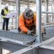 Installers fit steel grating panels and secure a guardrail post while building an elevated catwalk inside a commercial structure.