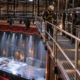 A lighting technician inspects a stage fixture from a guarded theatre catwalk while an engineer checks the rigging access route.