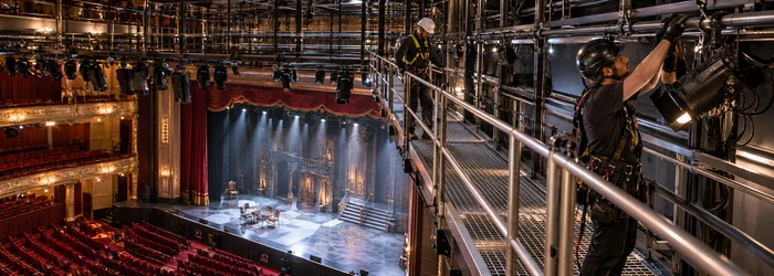 A lighting technician inspects a stage fixture from a guarded theatre catwalk while an engineer checks the rigging access route.