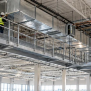 A maintenance worker inspects ceiling ductwork from a guarded steel catwalk with grating, support brackets, and stair access.