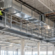 A maintenance worker inspects ceiling ductwork from a guarded steel catwalk with grating, support brackets, and stair access.