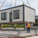 Crane sets a prefabricated modular office building beside site crews preparing connections.