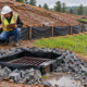 A site inspector checks a silt fence, protected storm drain, and stabilized drainage path at a construction site.