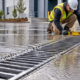 A worker checks a linear trench drain grate as rainwater flows across a paved loading area.