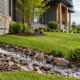 A downspout sends roof runoff into a rock-lined swale while an engineer checks the slope away from the house.
