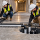 A civil engineer inspects an interior perimeter drainage channel leading to an accessible sump basin in a below-grade concrete building.
