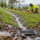A landscape crew checks a rock-lined swale and French drain that route runoff down a sloped yard into a catch basin.