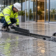 Installers inspect a grated linear drain across a wet commercial entrance forecourt to capture runoff before it reaches the doorway.