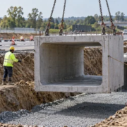 A crane lowers a precast concrete box culvert into a gravel-bedded road trench while an engineer checks alignment.