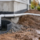 A technician checks an exterior sump basin as its discharge pipe carries water away from a foundation to a gravel swale.