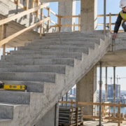 A carpenter checks tread and riser alignment on a cast-in-place concrete staircase while an engineer reviews the landing connection.