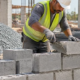 Mason lays concrete blocks on site beside staged cement, aggregate, reinforcing steel, and timber.