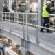 A maintenance technician inspects a valve from a guarded steel catwalk with grating beside industrial pipes.