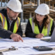 Construction estimator and project manager compare building plans, trade quotes, and a cost breakdown worksheet at an active site