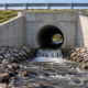 Concrete culvert headwall and wing walls directing water into a stone-lined channel as a civil engineer checks the outlet.