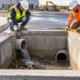 Construction workers inspect an open concrete catch basin and its stormwater inlet and outlet pipes.