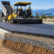 Highway construction crew inspects compacted asphalt layers and roadside drainage beside a paver and roller