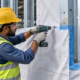 Worker fastens building wrap to steel framing and tapes overlapping seams to protect the structure from moisture