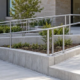 A wheelchair user follows a guarded switchback entrance ramp with level landing and continuous handrails while contractors review the route.