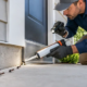 A gloved technician seals a small foundation gap where a trail of ants approaches a newly finished home.
