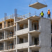 Mid-rise apartment building under construction with repetitive concrete floor slabs, open balcony bays, a tower crane, and workers.