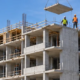 Mid-rise apartment building under construction with repetitive concrete floor slabs, open balcony bays, a tower crane, and workers.