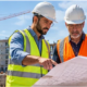 Apartment complex builder and project manager review construction plans in front of multi-unit residential buildings under construction.