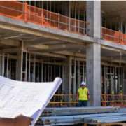 Architect in safety gear reviews a building drawing beside an active construction frame to check how the design is being built.