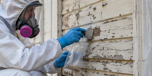 Environmental safety worker in protective clothing inspects aging exterior siding on a vintage home in a controlled work area.