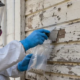 Environmental safety worker in protective clothing inspects aging exterior siding on a vintage home in a controlled work area.