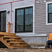 Builders finish siding and windows on the rear of a house under construction, with its deck and unfinished yard visible.