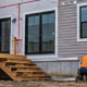 Builders finish siding and windows on the rear of a house under construction, with its deck and unfinished yard visible.