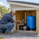 Insulated well house protecting a domestic pump and pressure tank from weather, with a technician checking the valve