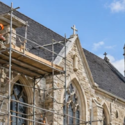 Masons restore a church facade and roof from scaffolding, carefully repairing stonework around the arched windows.