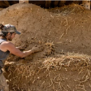Builder shapes a thick earthen cob wall mixed with visible straw while constructing a small natural-material house.