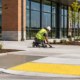 Workers finish a curb ramp and tactile warning surface along a sidewalk beside a new commercial building.