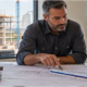 Commercial estimator and project manager review a building plan and scale ruler overlooking an active construction site, with no costs shown.