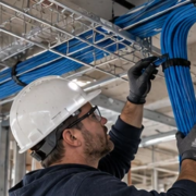 Electrician routes organized blue Ethernet cable through a tray above an unfinished commercial ceiling beside a network rack.
