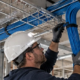 Electrician routes organized blue Ethernet cable through a tray above an unfinished commercial ceiling beside a network rack.