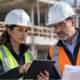 Field surveyor and architect review site measurements and building plans beside an unfinished structure, connecting fieldwork with design decisions.