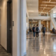 Long narrow corridor with doors and ceiling lights opening into a wider public hall with windows, seating, and gathering space.