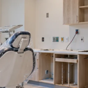 Dental operatory during construction fit-out, with a dental chair, treatment light, utility connections, cabinetry, and a contractor finishing the room.