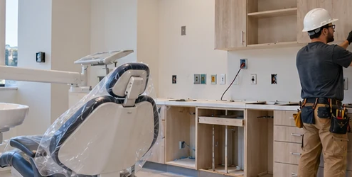 Dental operatory during construction fit-out, with a dental chair, treatment light, utility connections, cabinetry, and a contractor finishing the room.