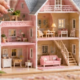 Hands place tiny furniture inside a pink multi-story miniature dream house being assembled on a craft table.