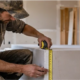 Drywall installer measures the edge of a gypsum board sheet in an unfinished residential room to check panel thickness before installation.