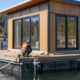 Modern floating house secured to a buoyant platform and moored on calm water as a crew aligns the deck and float system.