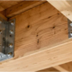Metal floor joist brackets secure intersecting timber joists to a support beam, with fastening holes visible.