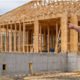 Homeowner and builder inspect a house mid-construction, showing a concrete foundation, timber frame, and roof trusses.
