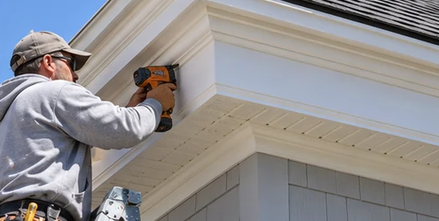 Craftsman installs projecting cornice trim where the roof eave meets the exterior wall, showing fascia, soffit, and weather-protective detailing.
