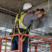 Worker secures suspended HVAC ducts and utility lines beneath a commercial floor slab using ceiling supports.